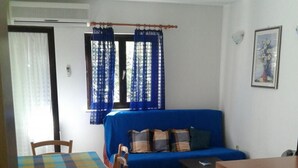 A compact living space with a blue sofa, a dining table with chairs, a small kitchenette, and a window with blue curtains. - Apartments Magda - Two Bedroom Apartment with Terrace and Garden View (Supetar)