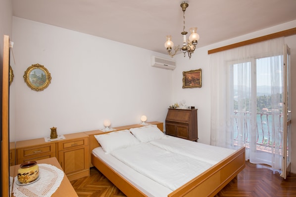 Une chambre à coucher avec un lit en bois, des tables de chevet, une commode et une vue sur la mer à travers de fines rideaux.