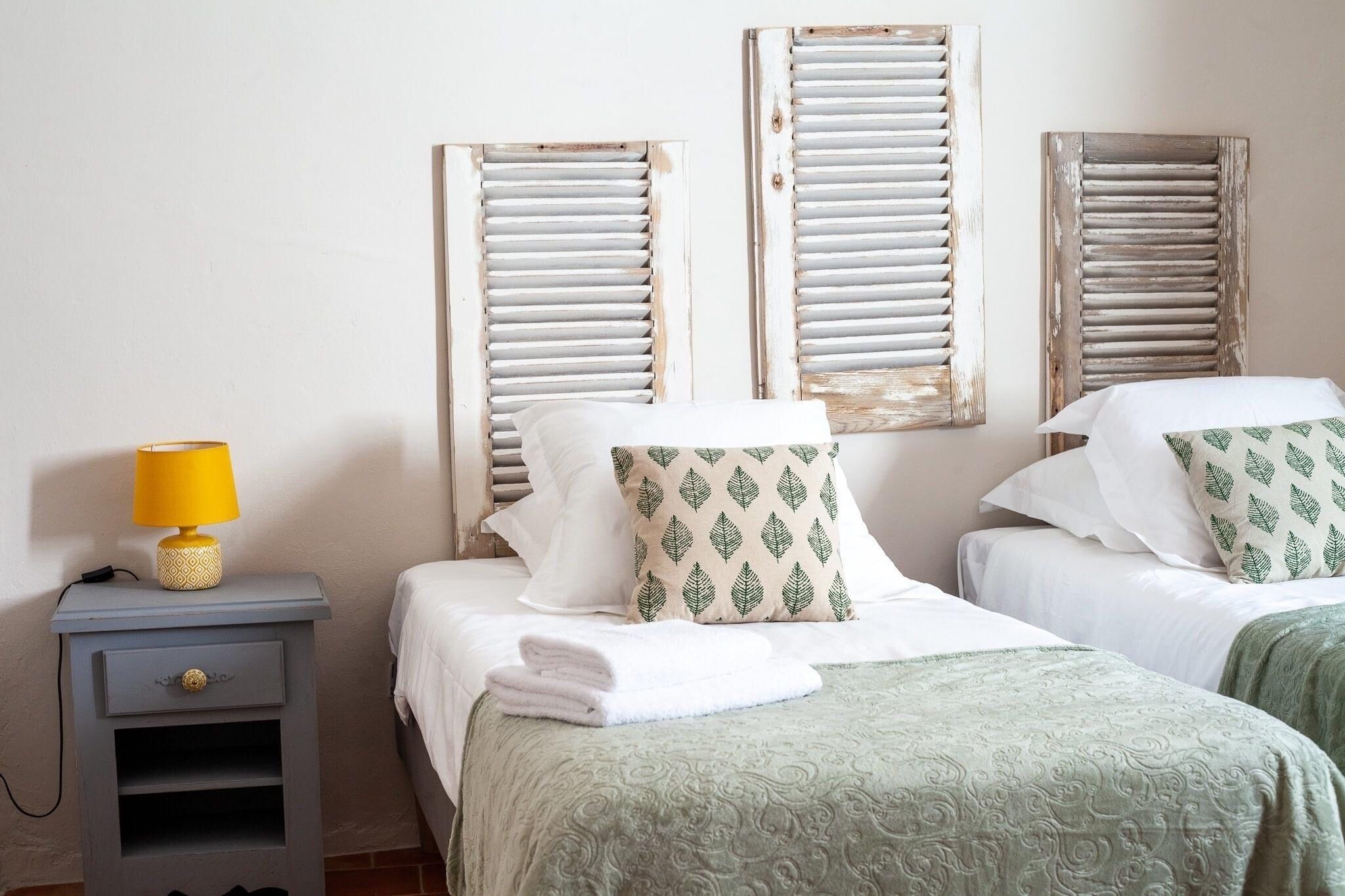 Un dormitorio con dos camas, una mesita de noche con lámpara y contraventanas en las ventanas.