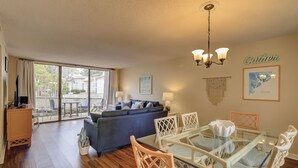 A spacious living area with a glass dining table, wicker chairs, a blue sofa, and a view of the outdoors through large sliding doors. - Lagoon Views & Steps to the Beach/Coligny Plaza! (Hilton Head Island)
