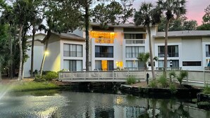 A two-story building with a balcony, a reflective water body in front, and surrounding trees. - Lagoon Views & Steps to the Beach/Coligny Plaza! (Hilton Head Island)
