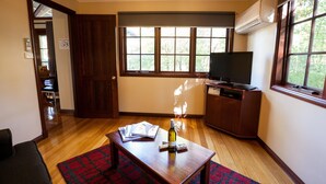 A room with a wooden floor, a red and black patterned rug, a wooden table with a bottle and magazines, a television on a wooden cabinet, and a window with a view of trees.
