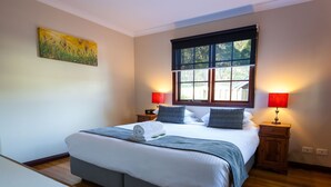 A neatly made bed with a blue and white bedspread, two bedside tables with lamps, a window with a view of greenery, and a painting on the wall.