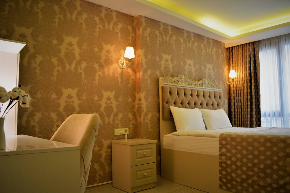 A hotel room with a tufted headboard, a bedside table with drawers, a lamp, and a large window with curtains.