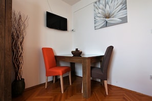 Un comedor con una mesa de madera, dos sillas, un jarrón con ramas, un cuadro de una flor y un televisor montado en la pared.