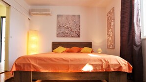 Una cama con estructura de madera y ropa de cama naranja, un aire acondicionado instalado en la pared, y un cuadro con flores.