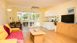 A living room with a sofa, a coffee table, and a TV. There is a kitchen area with cabinets and a plant. - Apartments Radic - Studio Apartment (2-3 Adults) - Kneza Domagoja Street - BR 3 (Dubrovnik)