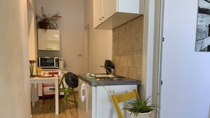 Eine kleine Küche mit Mikrowelle, Kaffeemaschine und Spüle. Zwei Stühle, einer grün, einer gelb, mit einem Topf mit Pflanze auf dem gelben Stuhl.