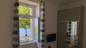 Ein Schlafzimmer mit großem Fenster, einem an der Wand befestigten Fernseher, einem Nachttisch mit Pflanze und einem Kleiderschrank.