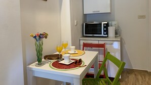 Eine kleine Küchenzeile mit einer Mikrowelle, einer Kaffeemaschine und einem weißen Schrank darüber. Ein Tisch mit einer Blumenvase, zwei Gläsern Orangensaft und einer Sonnenbrille auf einer Tischmatte. Ein grüner Stuhl steht an dem Tisch.