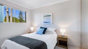 Un dormitorio con cama, dos mesitas de noche, un ventanal con vista a árboles y cielo, y un cuadro en la pared.