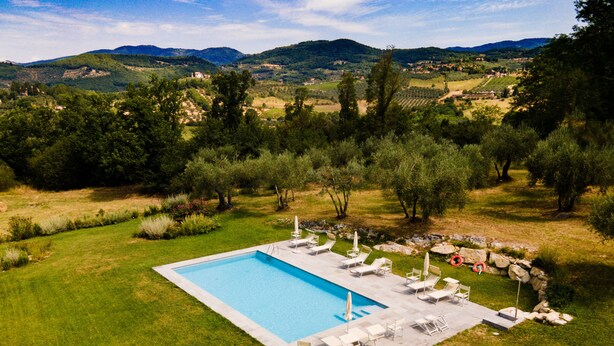 A rectangular swimming pool with a clear blue water surface, surrounded by a stone border. There are multiple white lounge chairs and umbrellas arranged around the pool. The pool is set in a well-maintained grassy area with various shrubs and trees in the background.