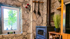 A rustic room with a stone wall, a window with a view of greenery, a wood-burning stove, and a shelf with logs and a potted plant. - Holiday house with private pool in a quiet location (Podašpilje)