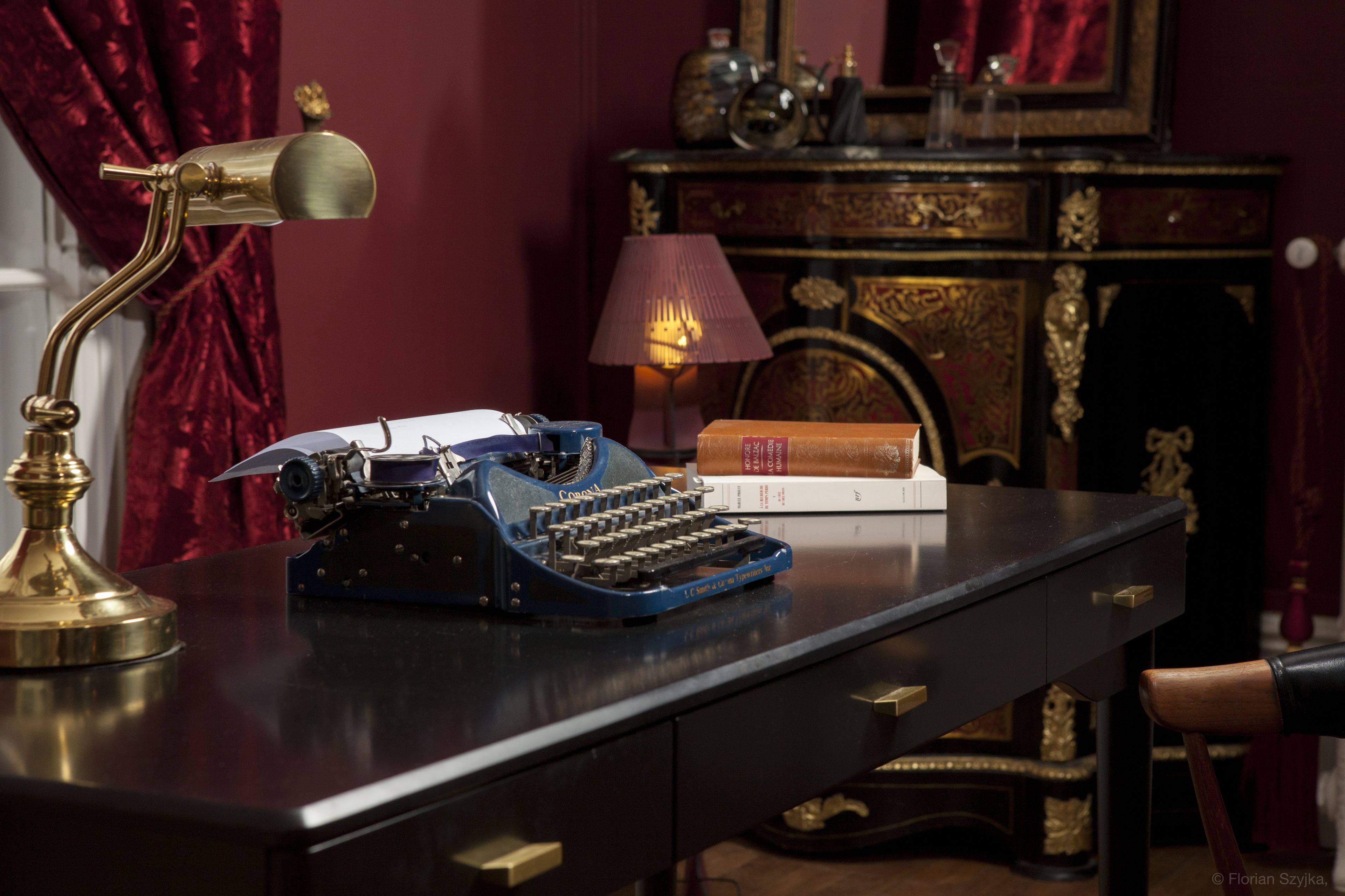 A vintage typewriter on a desk with a brass lamp, books, and a fireplace in the background.