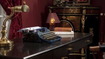 A vintage typewriter on a desk with a brass lamp, books, and a fireplace in the background.