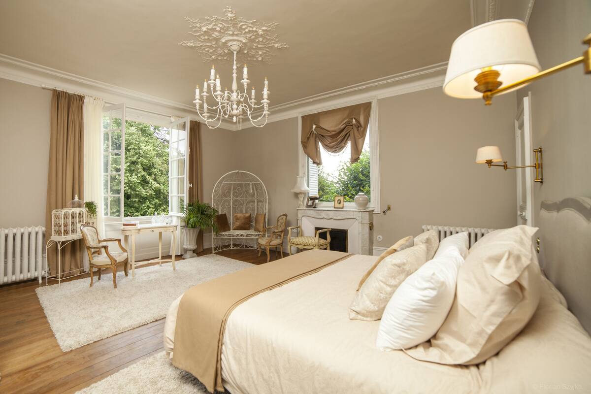Een slaapkamer met een groot bed, een kroonluchter, een bureau met stoel, en uitzicht op groen buiten.