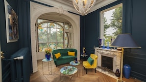 A room with a green sofa, yellow armchair, and a fireplace. There is a large window with stained glass, a chandelier, and a blue vase on a side table. - Villa Alecya (Sainte-Catherine-de-Fierbois)