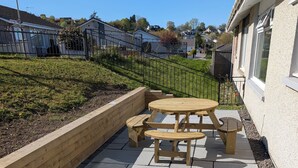 Une terrasse avec une table en bois et des bancs, entourée de dalles de pierre et d’un petit muret en briques.