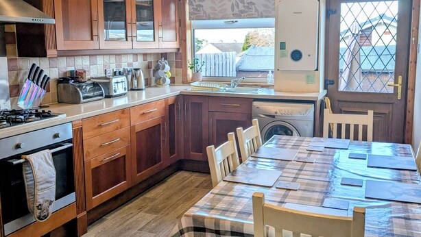 Une cuisine avec des meubles en bois, une table à manger avec des chaises, une cuisinière, un évier et une fenêtre donnant sur l’extérieur.