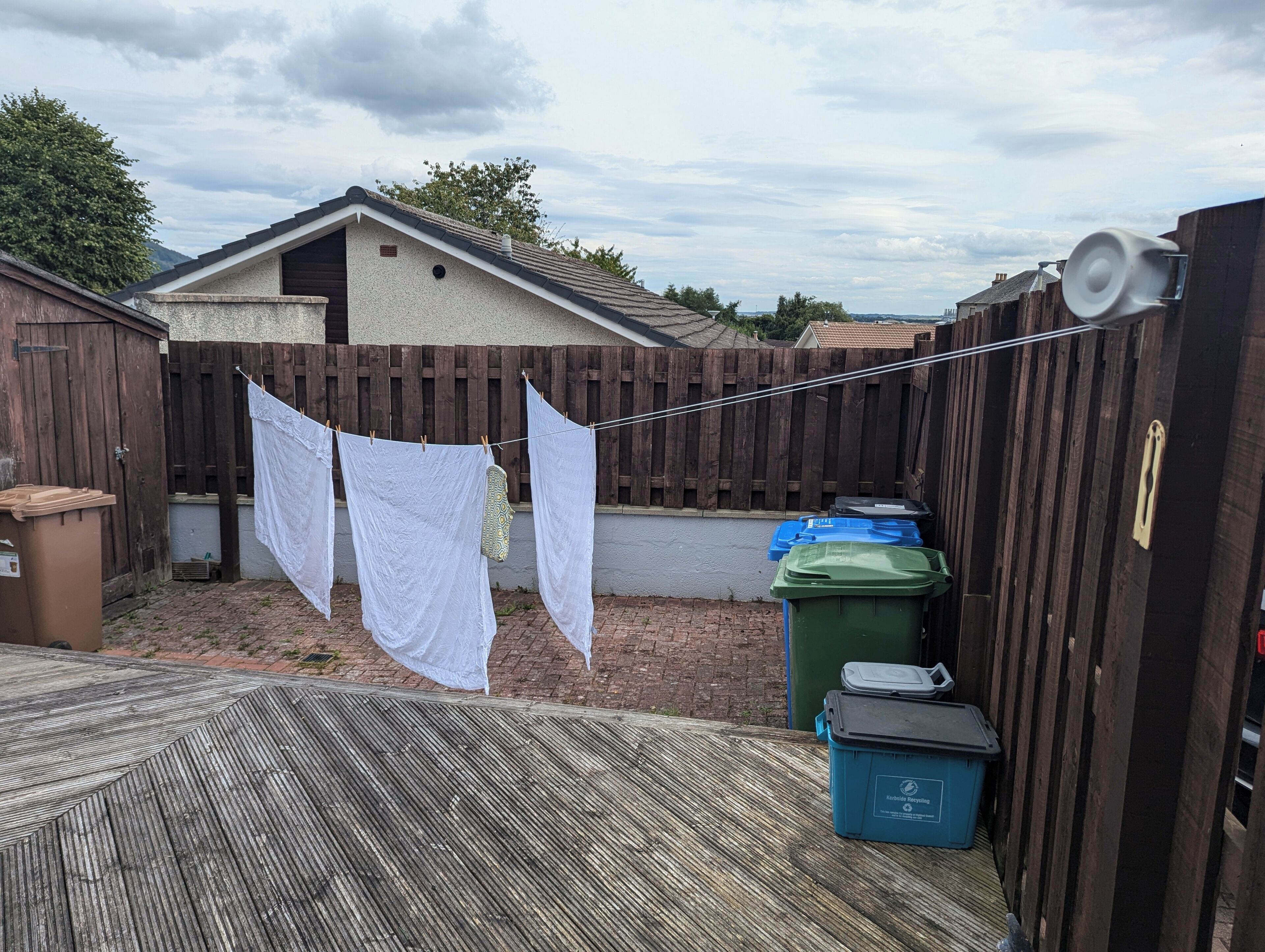 Une cour arrière avec une clôture en bois, un fil à linge où sont suspendus des vêtements blancs et des bacs de recyclage.