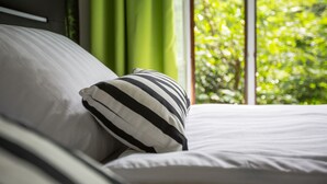 A bed with white bedding and a striped pillow, a window with green curtains, and a view of greenery outside. - "Waldblick" - 90 m² oasis of tranquility: Modern design & 2 bathrooms (Bad Sachsa)