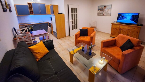 Un salon comprenant un canapé, deux fauteuils, une table basse, un téléviseur et un coin cuisine avec des placards et un réfrigérateur.