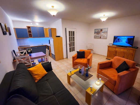 Un salon comprenant un canapé, deux fauteuils, une table basse, un téléviseur et un coin cuisine avec des placards et un réfrigérateur.
