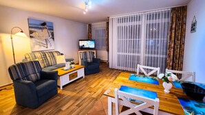 Een woonkamer met een bank, fauteuil, televisie, houten vloer en een eettafel met stoelen.