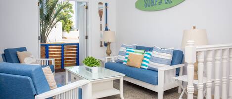 Una habitación con un sofá azul, sillones blancos, una mesa de centro de vidrio y un letrero en la pared que dice 'SURFS UP'.