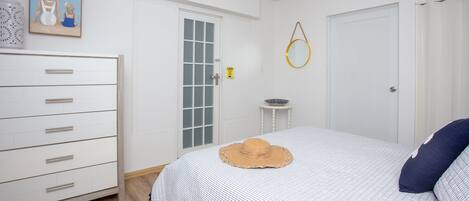 Ein Schlafzimmer mit Bett, Kommode, Tür, einem kleinen Tisch und einem gerahmten Bild an der Wand.