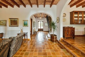 A spacious hallway with a tiled floor, wooden beams, a staircase, and a view into another room.