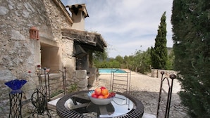 Ein Steingebäude mit Pool im Hintergrund, ein Tisch mit Obstschale und ein Metallstuhl.