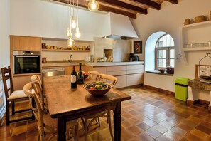 Een keuken met een houten tafel, stoelen, een schaal groenten en een wijnfles.