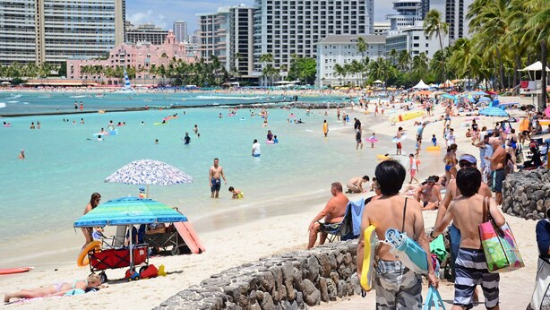 A crowded beach with people swimming and sunbathing, a lifeguard station, and tall buildings in the background.