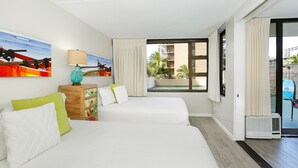 A hotel room with two beds, a window with a view of a building and palm trees, a small table with a lamp, and a wall-mounted painting of a boat.