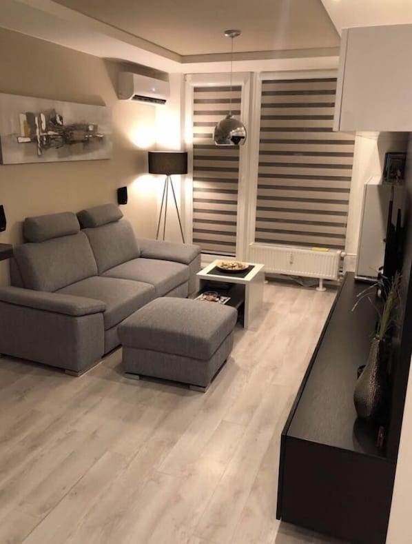 A modern living room with a grey sofa, a small ottoman, a floor lamp, and a radiator.