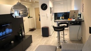 Un salon moderne avec un téléviseur à écran plat, un canapé et une table basse. Il y a un coin cuisine avec une table à manger et des chaises, ainsi qu’un petit coin bar avec un ordinateur portable et une bouteille.