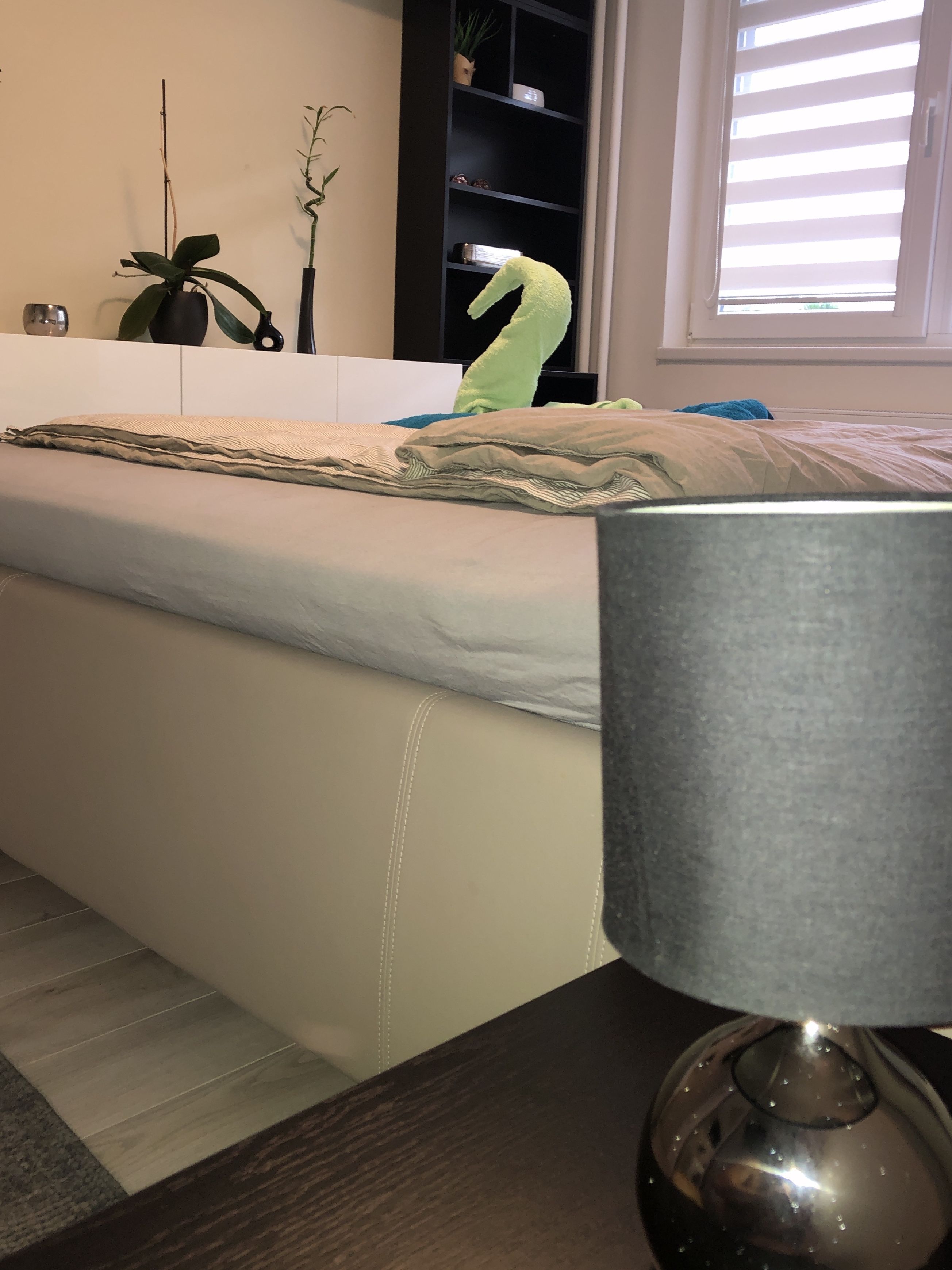 Una cama con colchón blanco y cabecero gris, una mesita de noche con lámpara y una ventana con persianas.