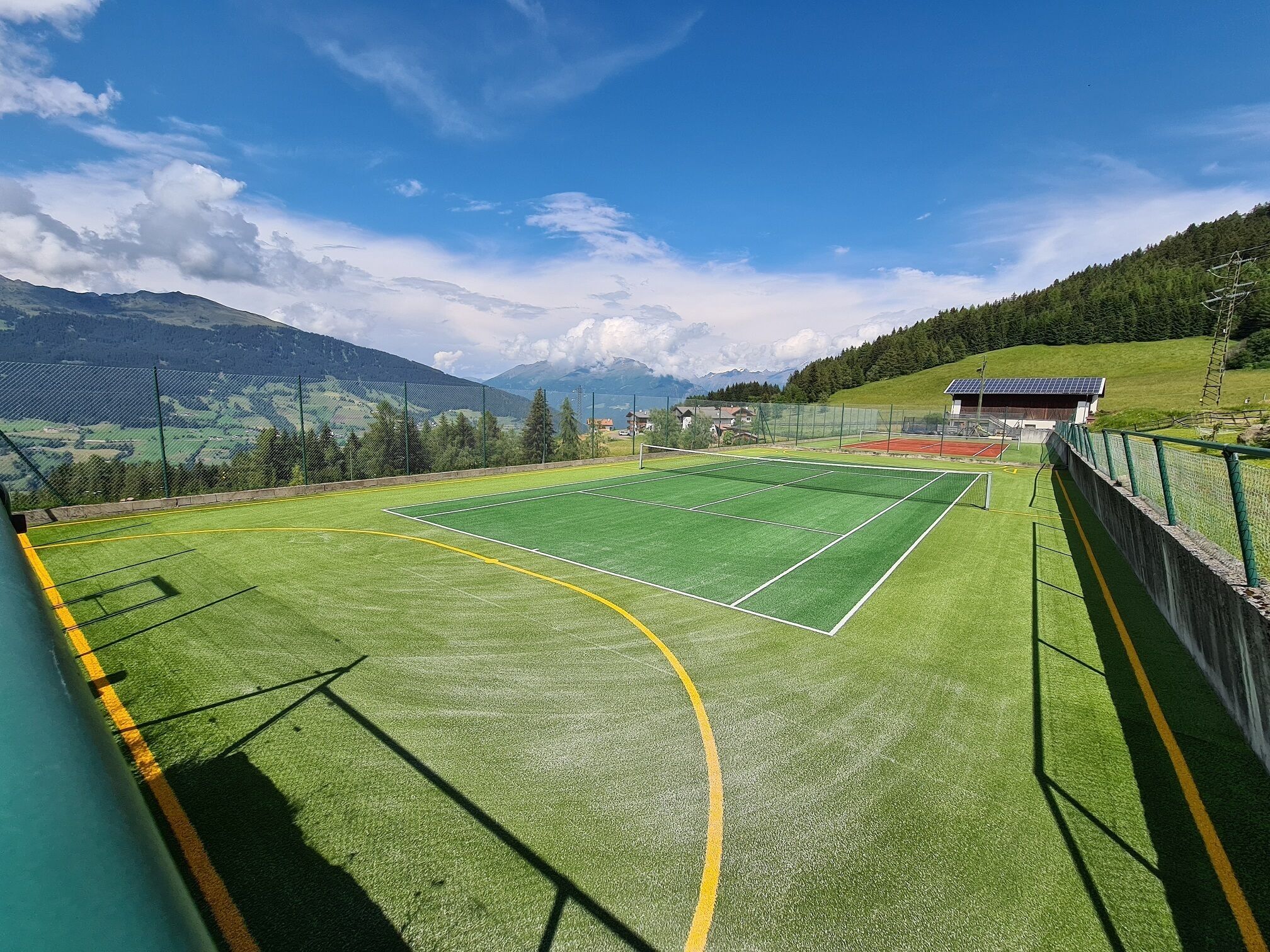 Ein Tennisplatz mit Netz, umgeben von einem Zaun, und ein Gebäude mit Solaranlage im Hintergrund.