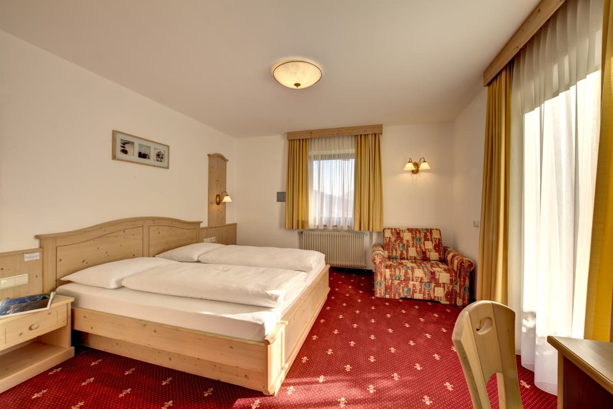 Une chambre d’hôtel avec un lit en bois, une moquette rouge à motifs, une chaise, un bureau et une fenêtre avec des rideaux.