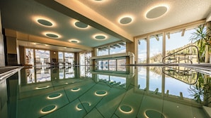 A spacious indoor swimming pool with multiple circular light fixtures on the ceiling, large windows offering views of the outdoors, and a glass railing. - Sporthotel Kalcherhof (Racines)