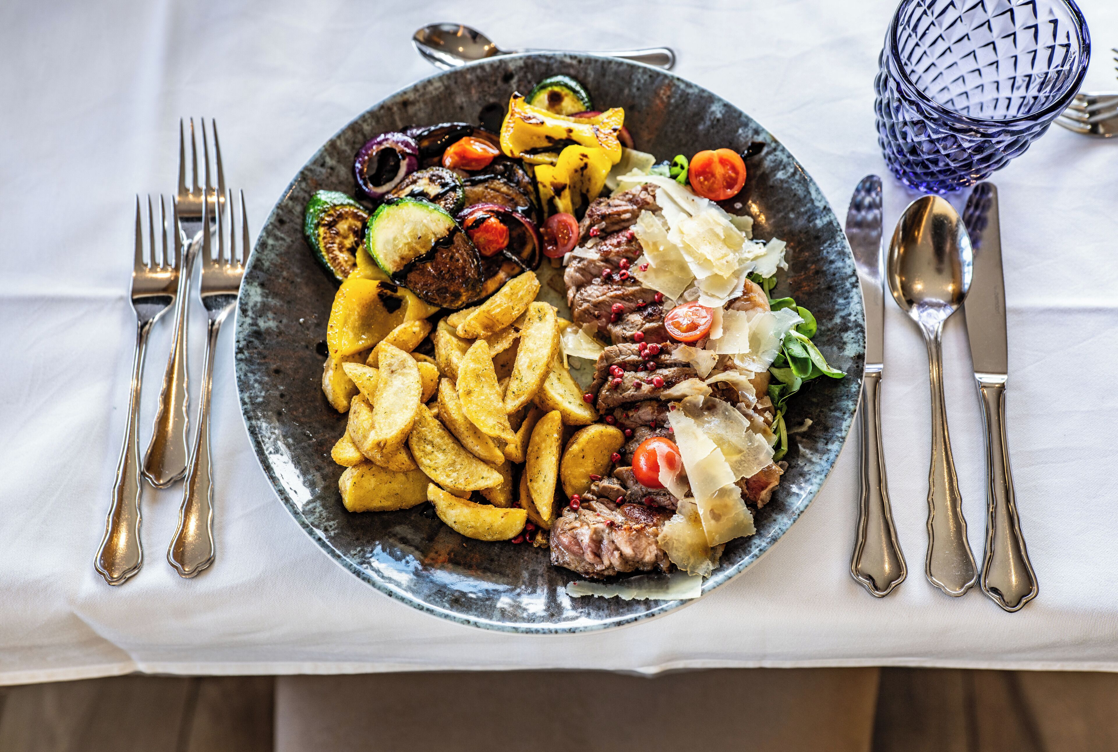 A plate of food with various vegetables and meat, accompanied by cutlery on a tablecloth.
