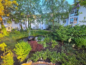 Ein Garten mit Teich, verschiedenen Pflanzen und einem Gebäude im Hintergrund.