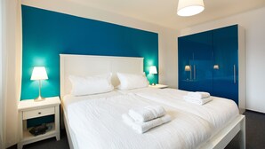 Ein ordentlich bezogenes Bett mit weißen Bettwäsche und Handtüchern, ein weißer Nachttisch mit Lampe und eine blaue Akzentwand.