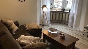 Un salon chaleureux avec un canapé marron foncé, une petite table basse en bois et un lampadaire. Une grande fenêtre ornée de rideaux blancs offre une vue sur le paysage urbain.