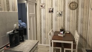 Une cuisine compacte équipée d’une machine à café, d’un évier et d’une table à manger avec des chaises. Une horloge est fixée au mur, et de petites photos encadrées y sont accrochées.