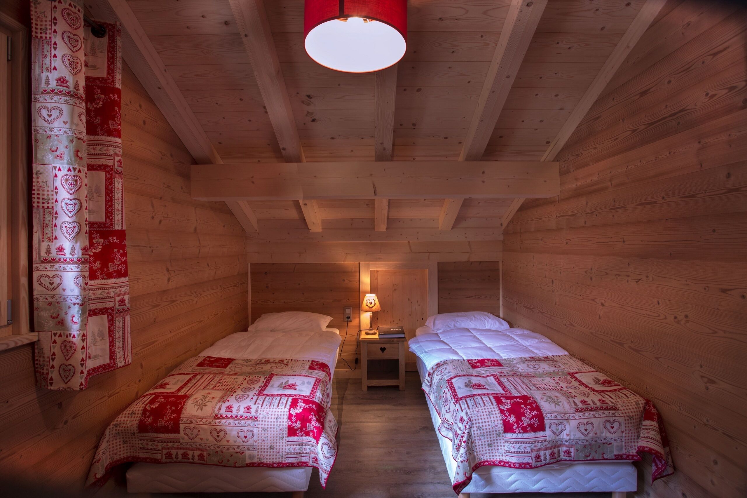 A cozy room with two beds, wooden walls, a red ceiling light, and a patterned curtain.