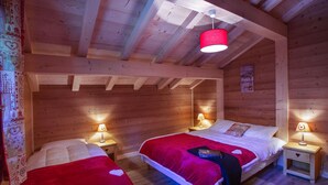 Une chambre chaleureuse en bois, aménagée comme un chalet, avec deux lits, chacun recouvert d’une couverture rouge et de draps blancs. On y trouve deux lampes de chevet et un luminaire rouge suspendu au plafond.