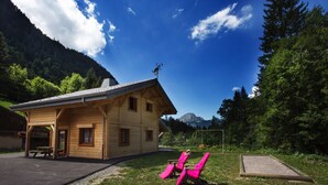 Une cabane en bois avec une table de pique-nique et deux chaises roses, située dans un espace verdoyant, entourée d’arbres et de montagnes.