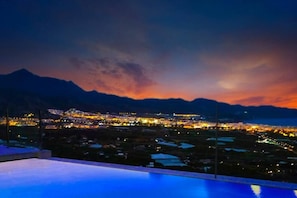 Une vue nocturne d’une ville, avec des montagnes en arrière-plan et une piscine au premier plan.